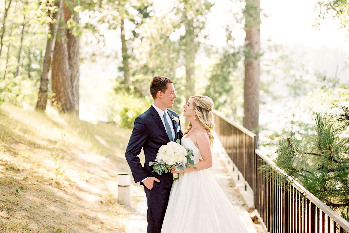 A couple in wedding attire stands on a wooded path with trees and sunlight filtering through, creating a romantic and serene scene.