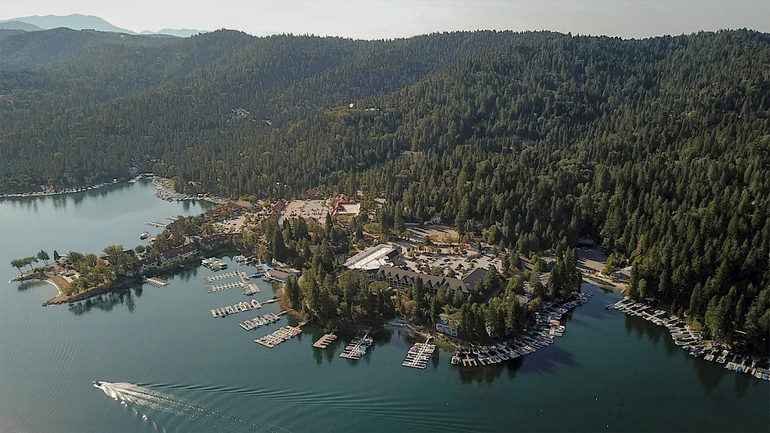 Aerial view of a forested lakeside marina village with boats, docks, and a dense tree line surrounding a calm blue lake.