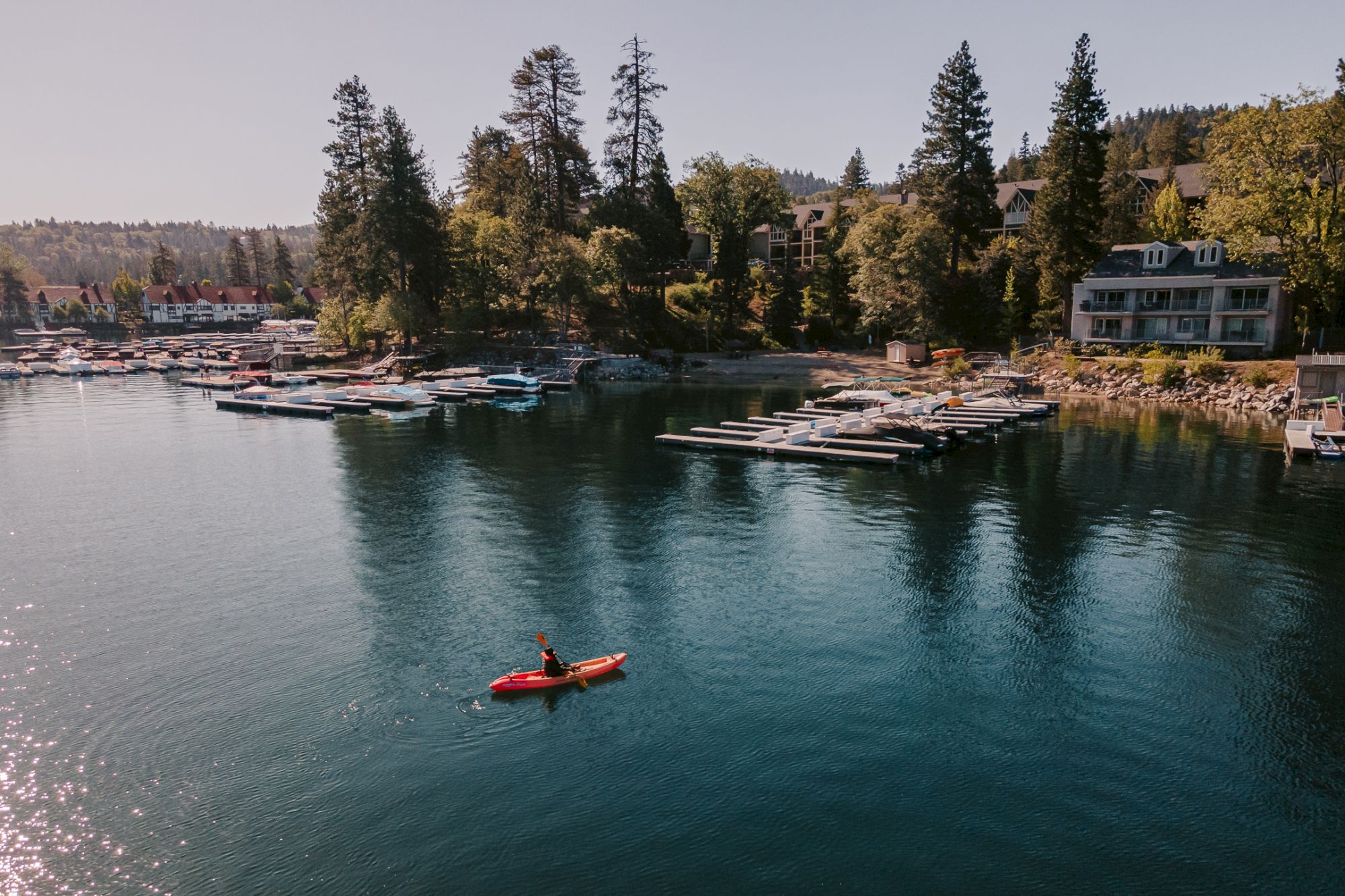 A calm lakeside marina with many docked boats and cabins, plus a red kayak paddler on the water.