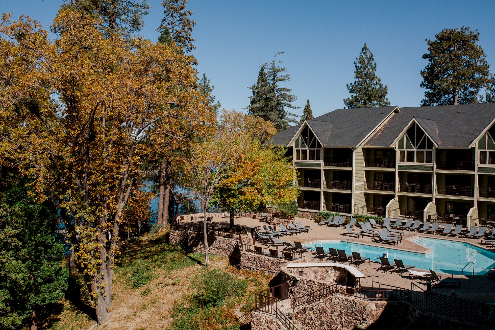 An outdoor hotel pool and lounge chairs beside a multi-story building, with autumn trees under a clear blue sky.