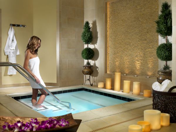 A woman in a towel uses the pool ladder in a cozy indoor spa with candles and neatly arranged decor.