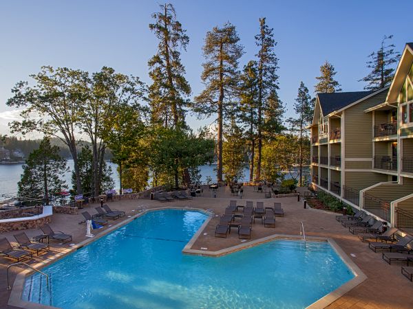 An outdoor hotel pool with lounge chairs by a lake, surrounded by tall trees and a lakeside building, under blue sky.