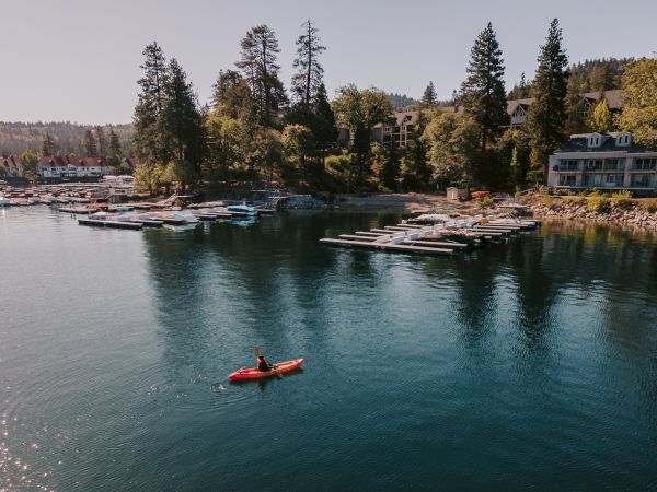 A calm lake marina with many docked boats and a red kayak, plus cabins and trees along the shore under a clear sky.