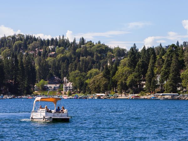 A small passenger boat cruises on a blue lake beside a marina, with lush forested hills and homes in the background.