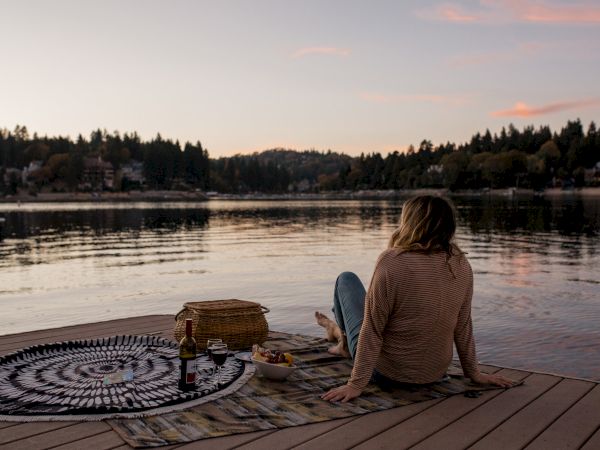 A person relaxes on a lakeside deck at sunset with a picnic, wine bottle, and drinks.