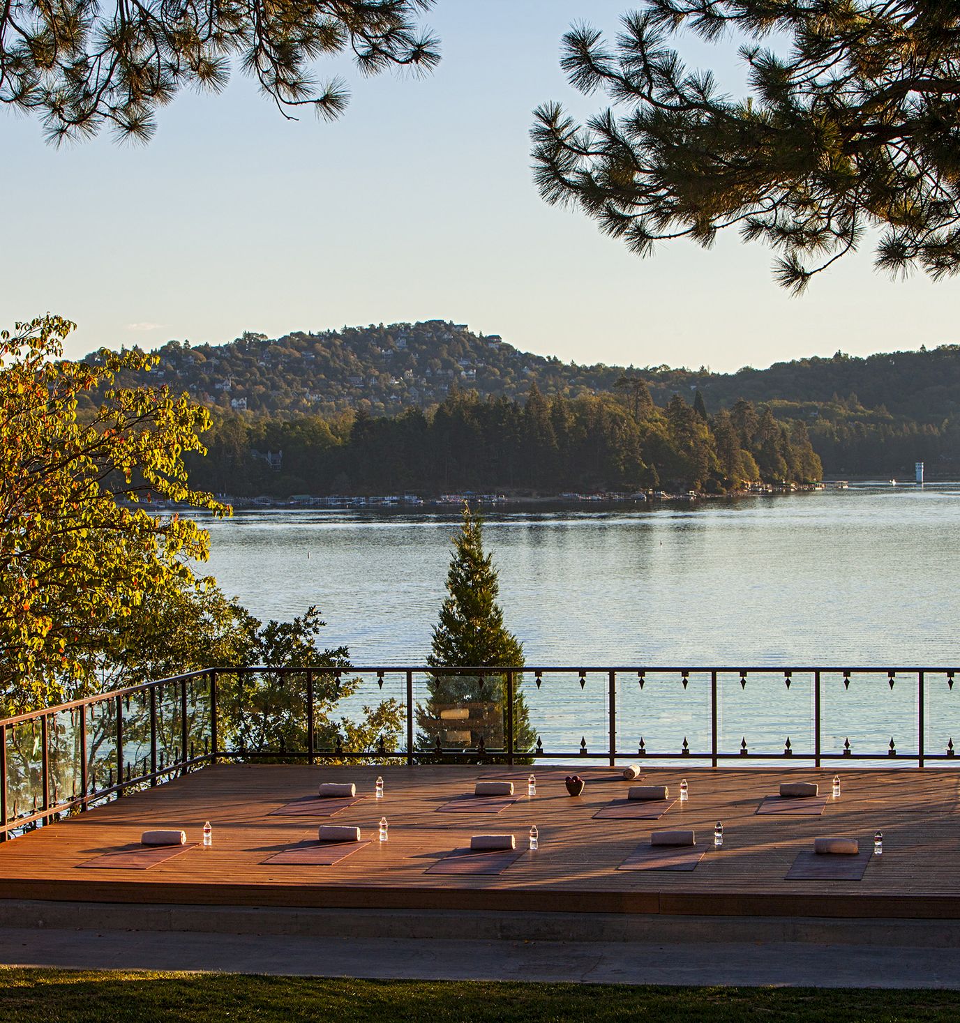 Sunlit outdoor deck with yoga mats and bottles overlooks a calm lake and wooded hills framed by pine trees.