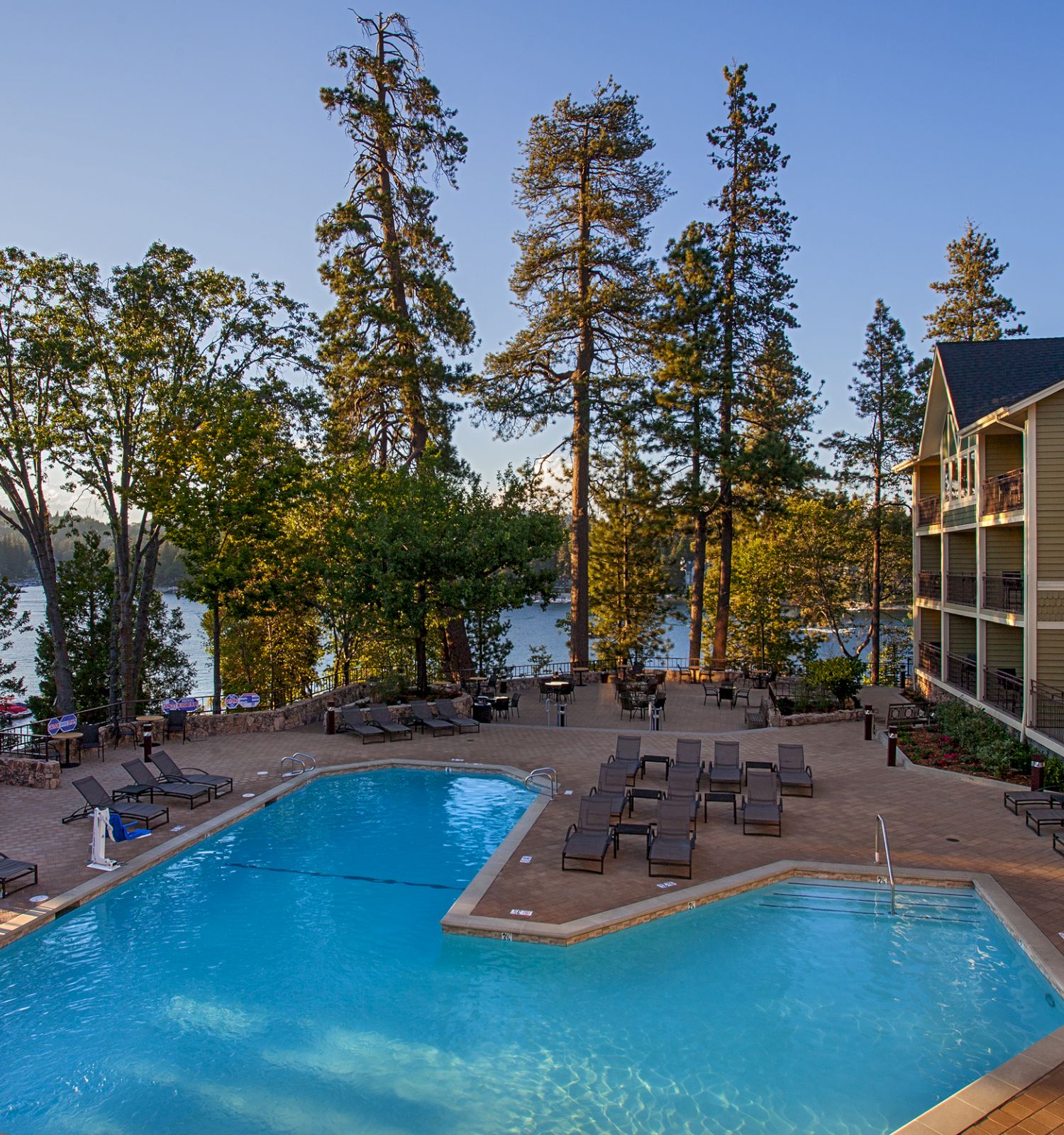 Outdoor hotel pool with lounge chairs and a view of a calm river, beside a multi-story building under a clear sky.