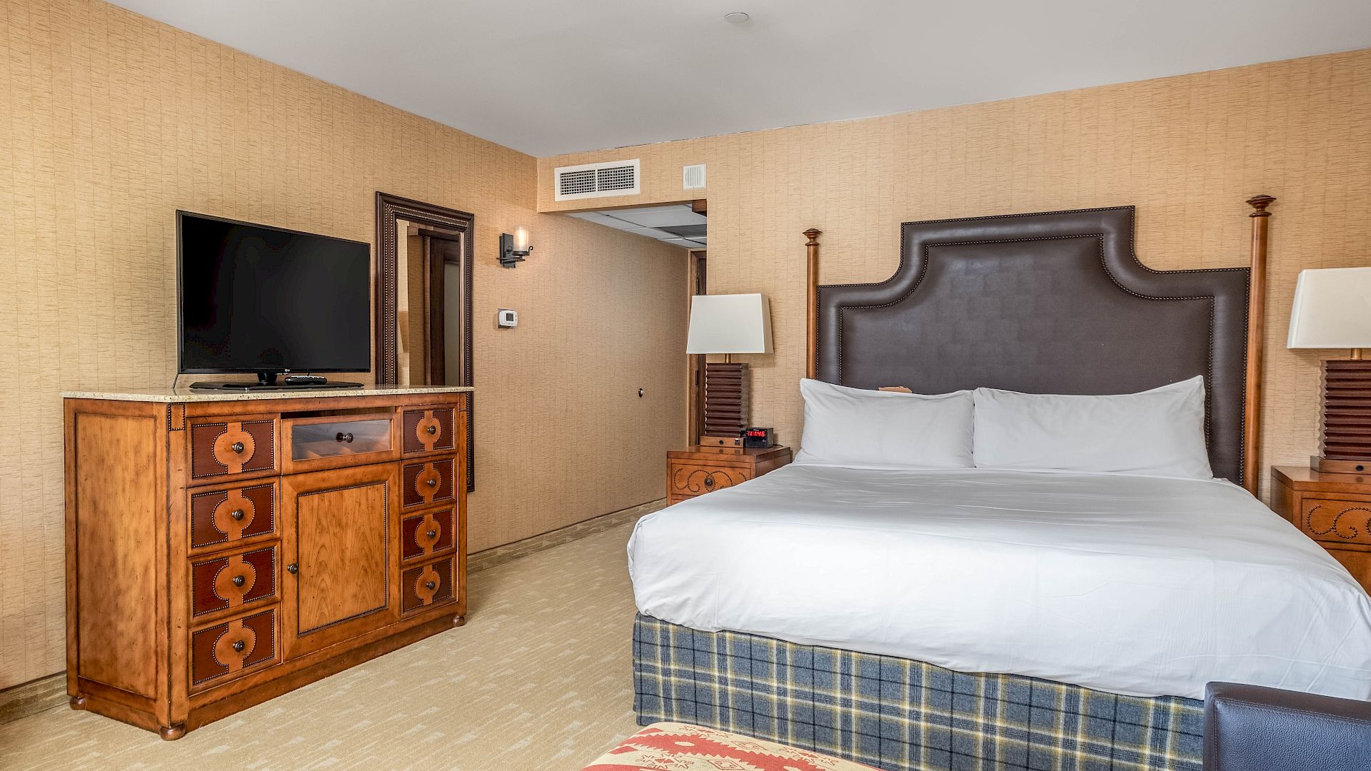 A hotel bedroom with a large TV on a wooden dresser, a bed with white linens, and bedside lamps against beige walls.