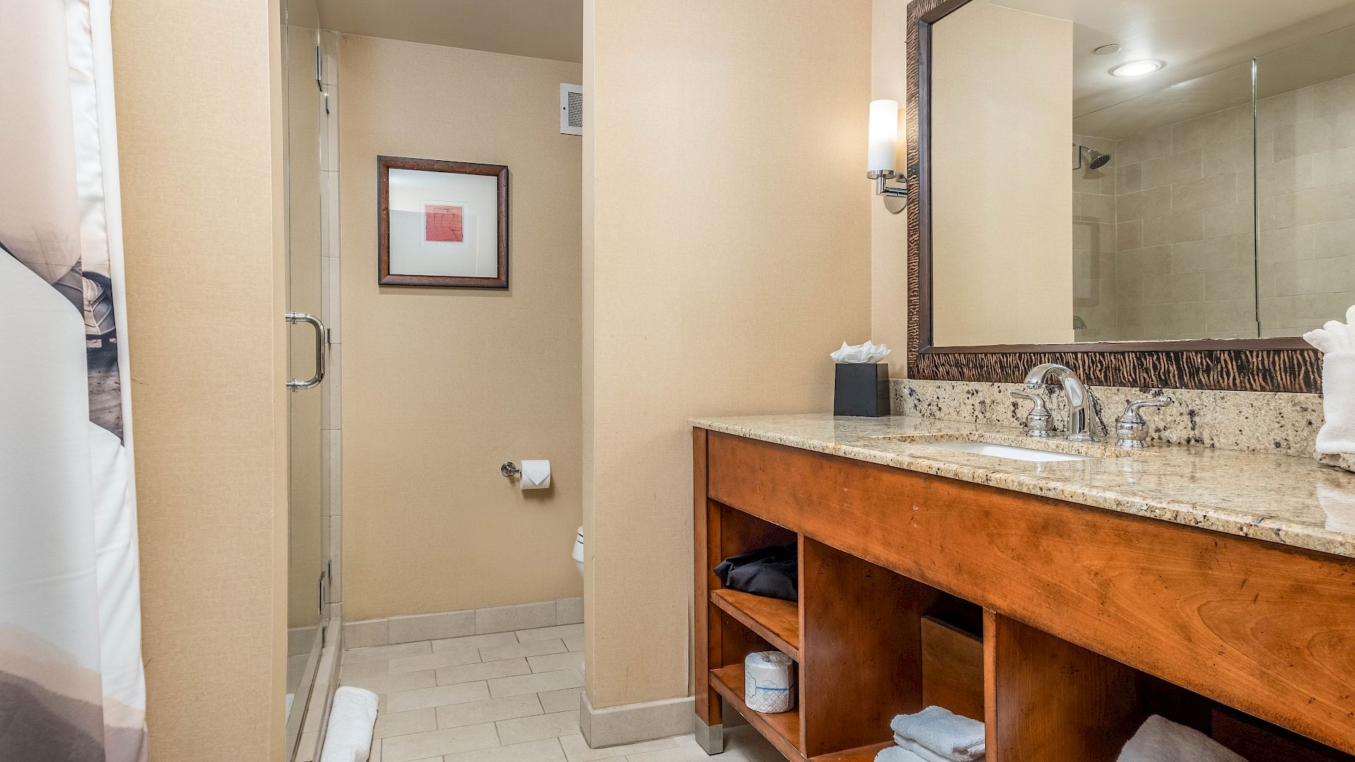 A modern hotel bathroom with a glass shower, granite vanity with faucets, towels, and a toilet tucked in the corner.