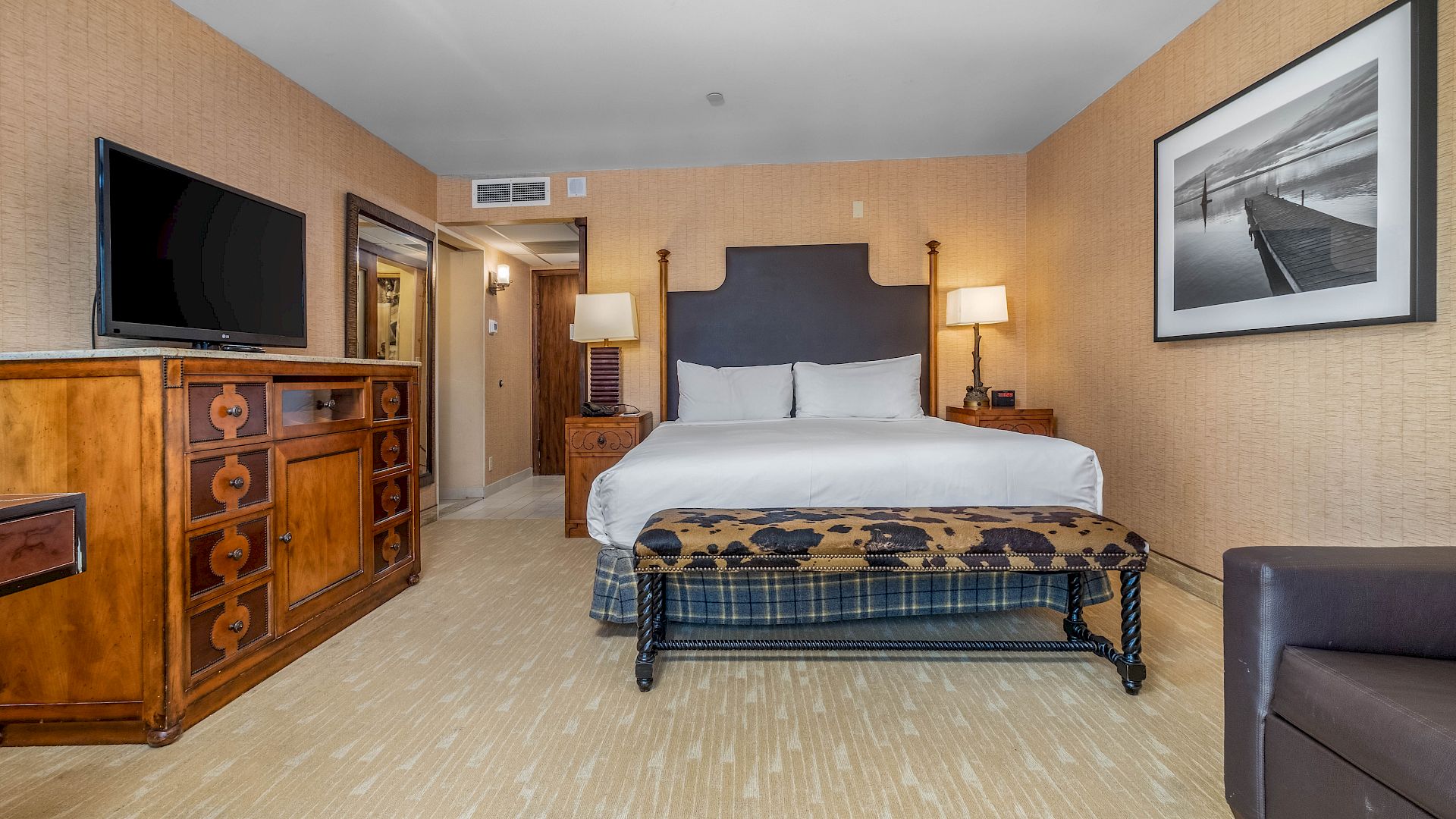 A hotel room with a large bed, dresser with TV, bedside lamps, and framed artwork on beige walls.