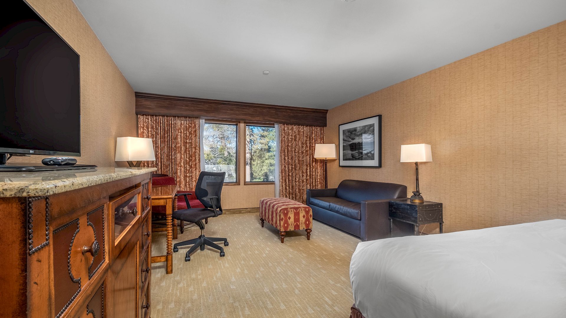 A tidy hotel room with TV and desk, swivel chair, blue couch, lamps, curtains, and a window view of trees.