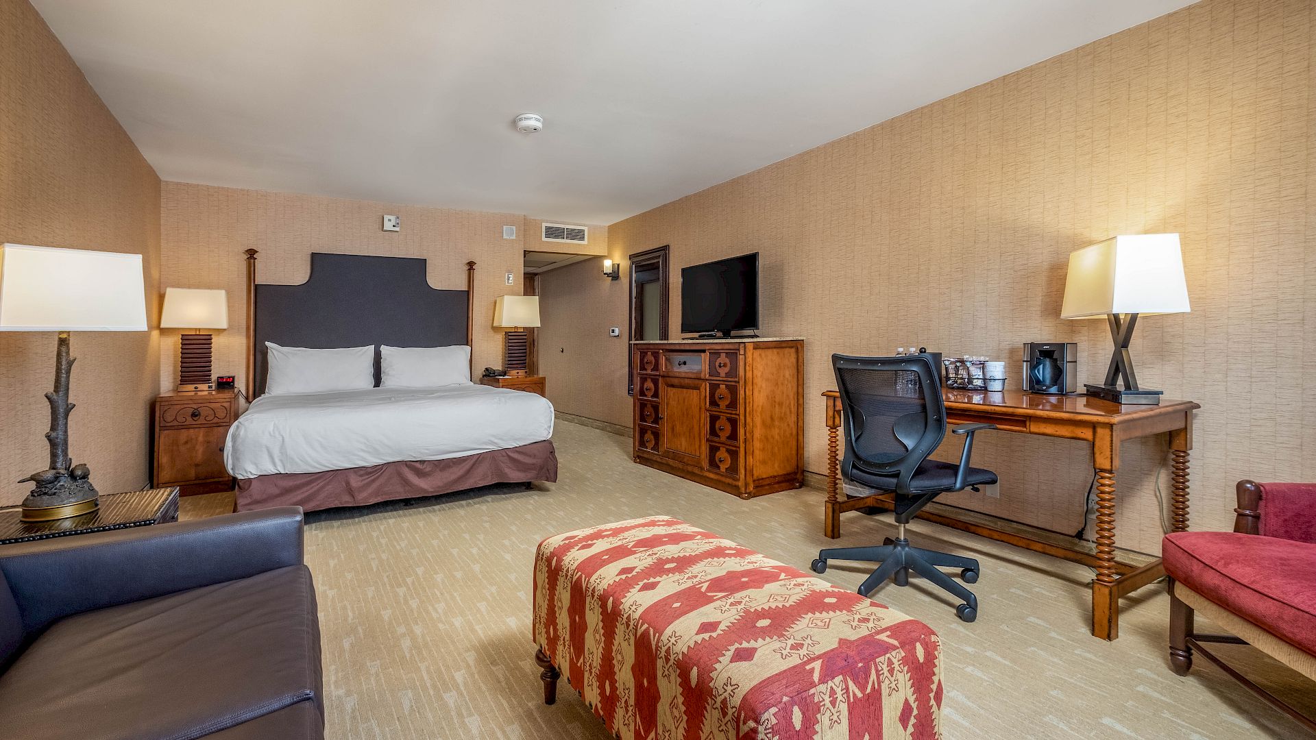 A cozy hotel suite with a king bed, TV entertainment center, desk coffee station, and lamps along the walls.
