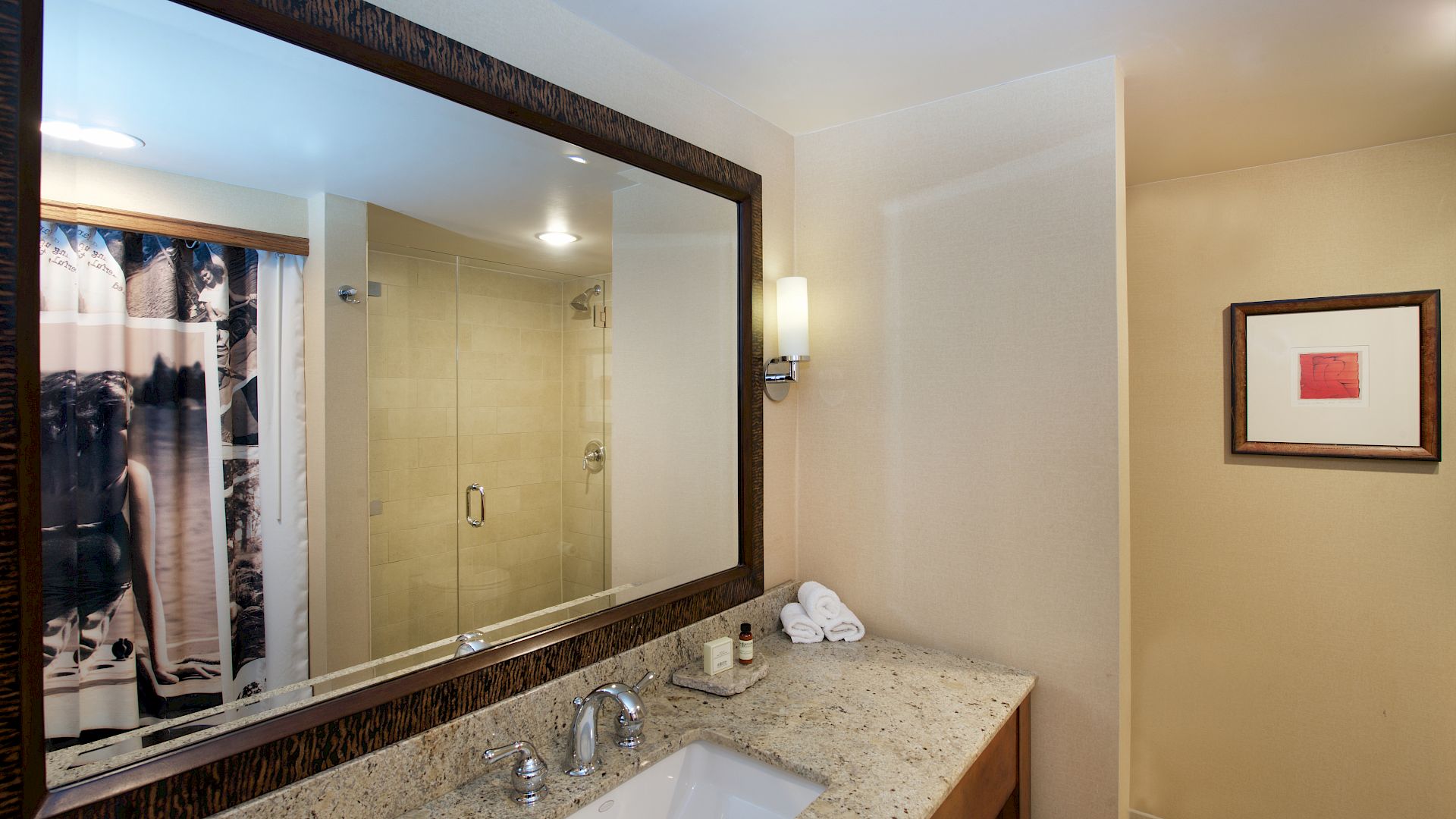 A hotel bathroom with a granite sink vanity, framed wall art, and a glass shower showing a patterned curtain and towels.