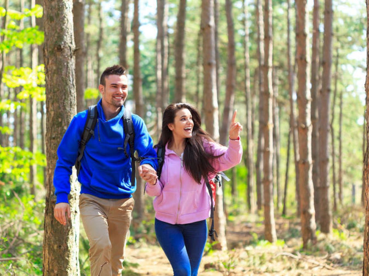 Two people are hiking in a sunny forest, smiling and enjoying the outdoors.