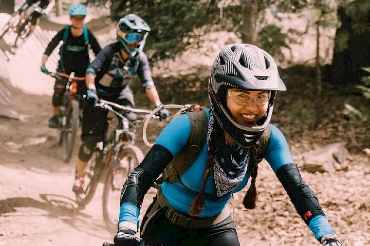 Cyclists with helmets ride mountain bikes on a dirt trail in a forested area, wearing sporty gear and backpacks, one smiling at the camera.