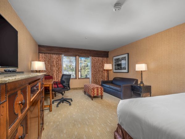 A hotel room office setup with a TV, desk, chair, sofa, lamps, framed art, curtains, and a window view.