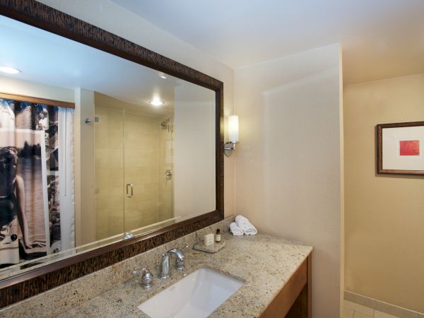 A hotel bathroom with a granite sink, framed art, and a walk-in shower with curtains, plus folded towels.