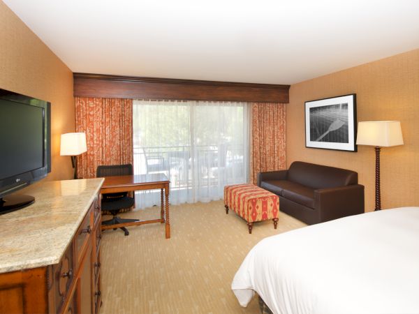 A hotel room with granite desk, flat-screen TV, curtains over a window, framed art, lamps, and a brown sofa-bed.