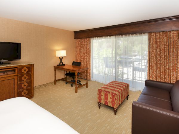 A cozy hotel-style bedroom with a TV, wooden desk and lamp, a bed, and large windows with patterned curtains and sheer drapes.