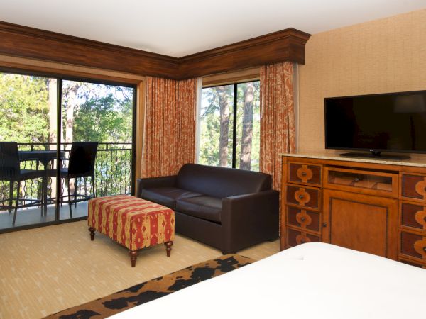 A cozy room with floor-to-ceiling windows, orange patterned curtains, a leather couch, a TV on a wooden dresser, and an outdoor view.