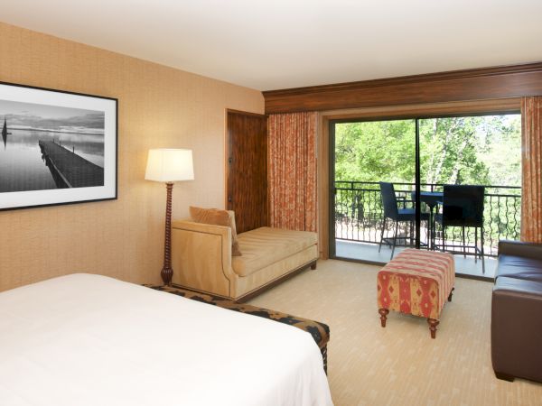 A hotel bedroom with a large bed, lamp, patterned bench, and a balcony door opening to green trees outside.