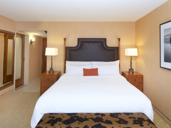 A hotel bedroom with a large bed, bedside tables and lamps, framed wall art, and warm beige walls.