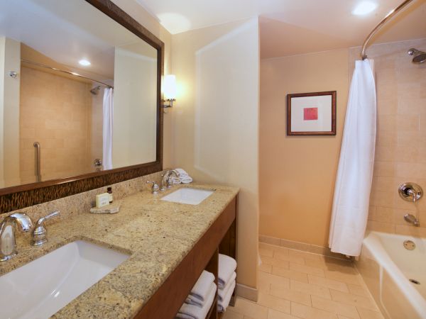 A bright hotel bathroom with granite double vanity, framed mirror, bathtub with showerhead, and white towels.