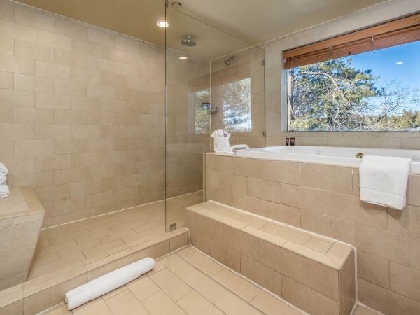A sunny tiled bathroom with a glass shower, rainfall head, bathtub, towels, and a window showing pine trees outside.