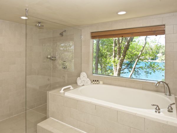 Bright bathroom with a glass shower, freestanding bathtub, folded towels, and a window showing trees and blue water.