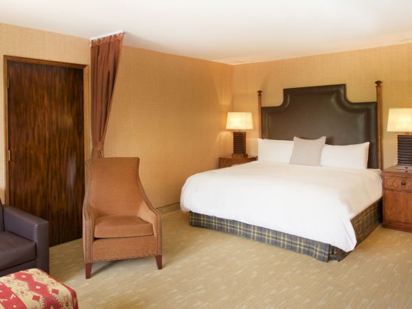 A cozy hotel bedroom with a king-size bed, nightstands, lamps, curtains, and seating by a wooden door.