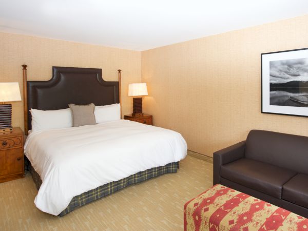 A neatly made hotel bedroom with a large dark headboard bed, bedside lamps, framed photo wall art, and a brown seating area.