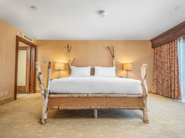 A cozy hotel bedroom with a king-size bed, lamps, birch-branch decor, warm walls, and patterned curtains by a window.