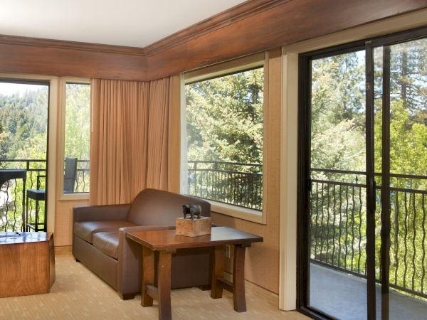 A cozy balcony living area with curtains, a couch and coffee table, and large windows showing a lush green forest view.