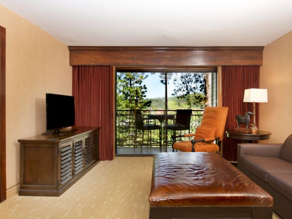 A hotel room lounge area with a leather sofa and bed, a TV console, and large windows showing chairs and green scenery outside.