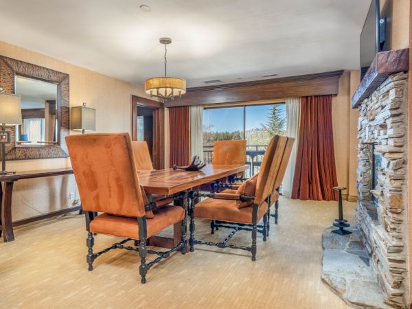 Bright dining room with a wooden table, leather chairs, chandelier, draped curtains, and a stone fireplace wall beside large windows.