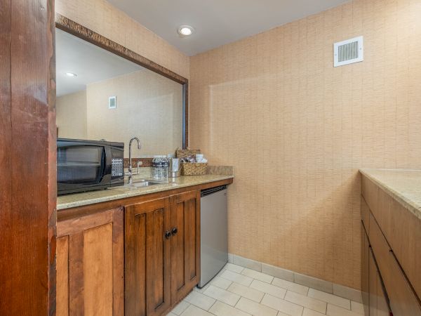 Small kitchenette with microwave, sink and faucet, granite counter, tan tile walls, and wicker baskets on top.