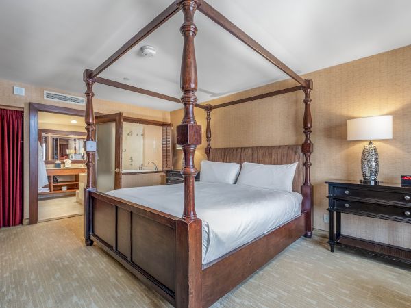 A four-poster bed in a hotel room, with white linens, side dresser, lamp, and a bathroom visible through the doorway.