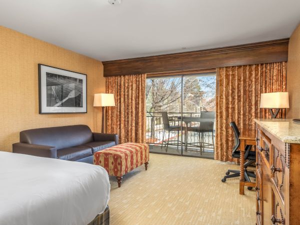 A cozy hotel room with a sofa, patterned curtains, a balcony view, lamps, and a TV on a wooden dresser.