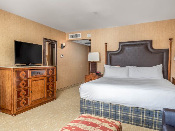 A hotel bedroom with a tufted headboard bed, white sheets, side tables, a large TV on a dresser, and beige wallpaper.