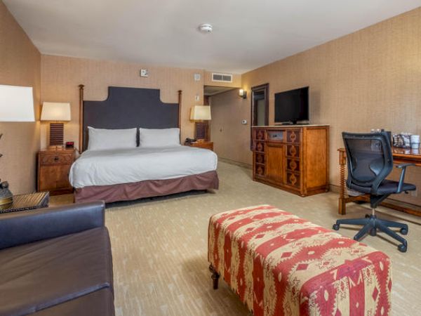 A hotel room with a large bed, patterned bench, desk with chair, and carpeted floor.