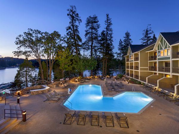 A lakeside hotel courtyard with two glowing pools, lounge chairs, and a multi-story building under a blue evening sky.