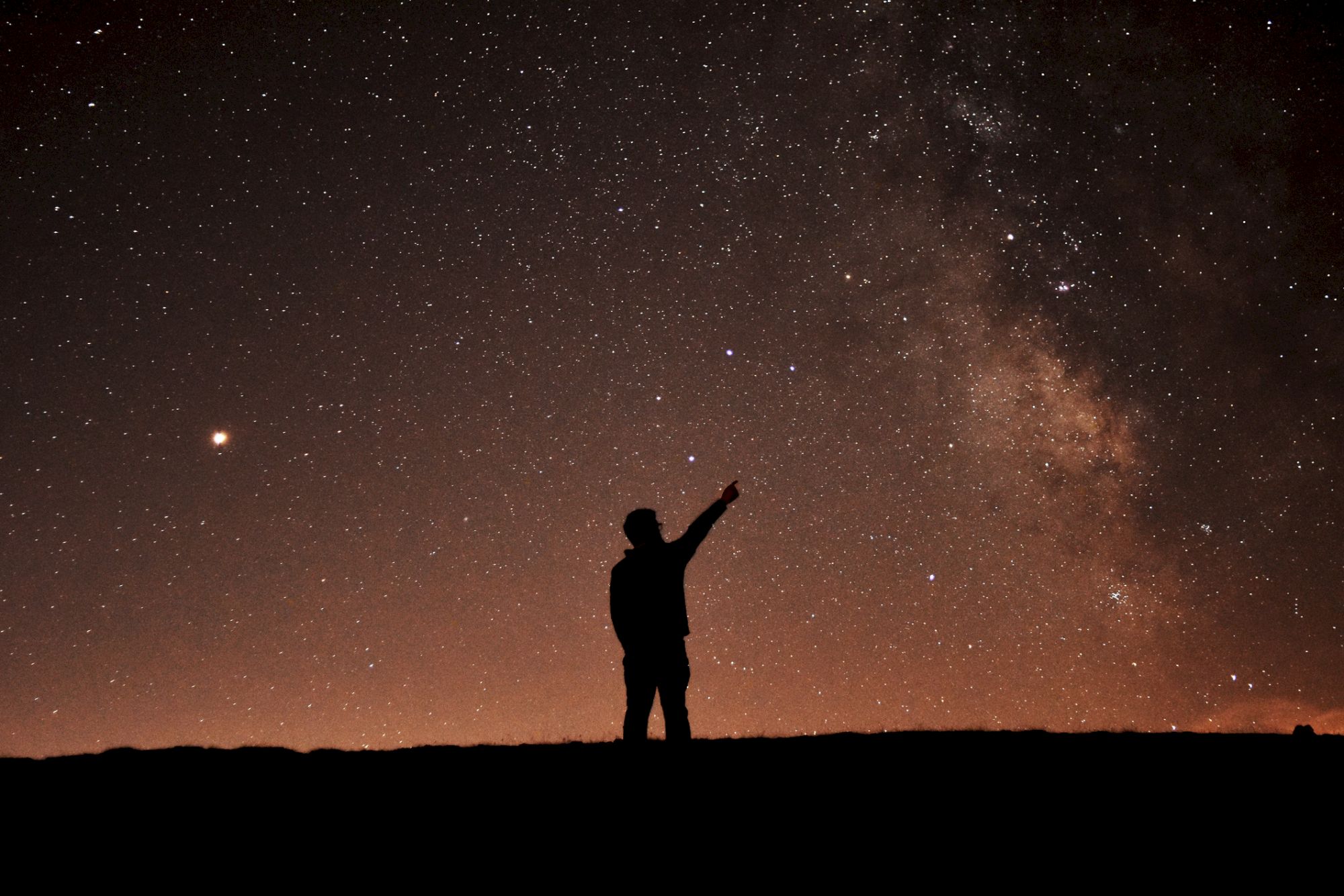 A silhouetted person points toward the bright Milky Way and scattered stars across a dark night sky.