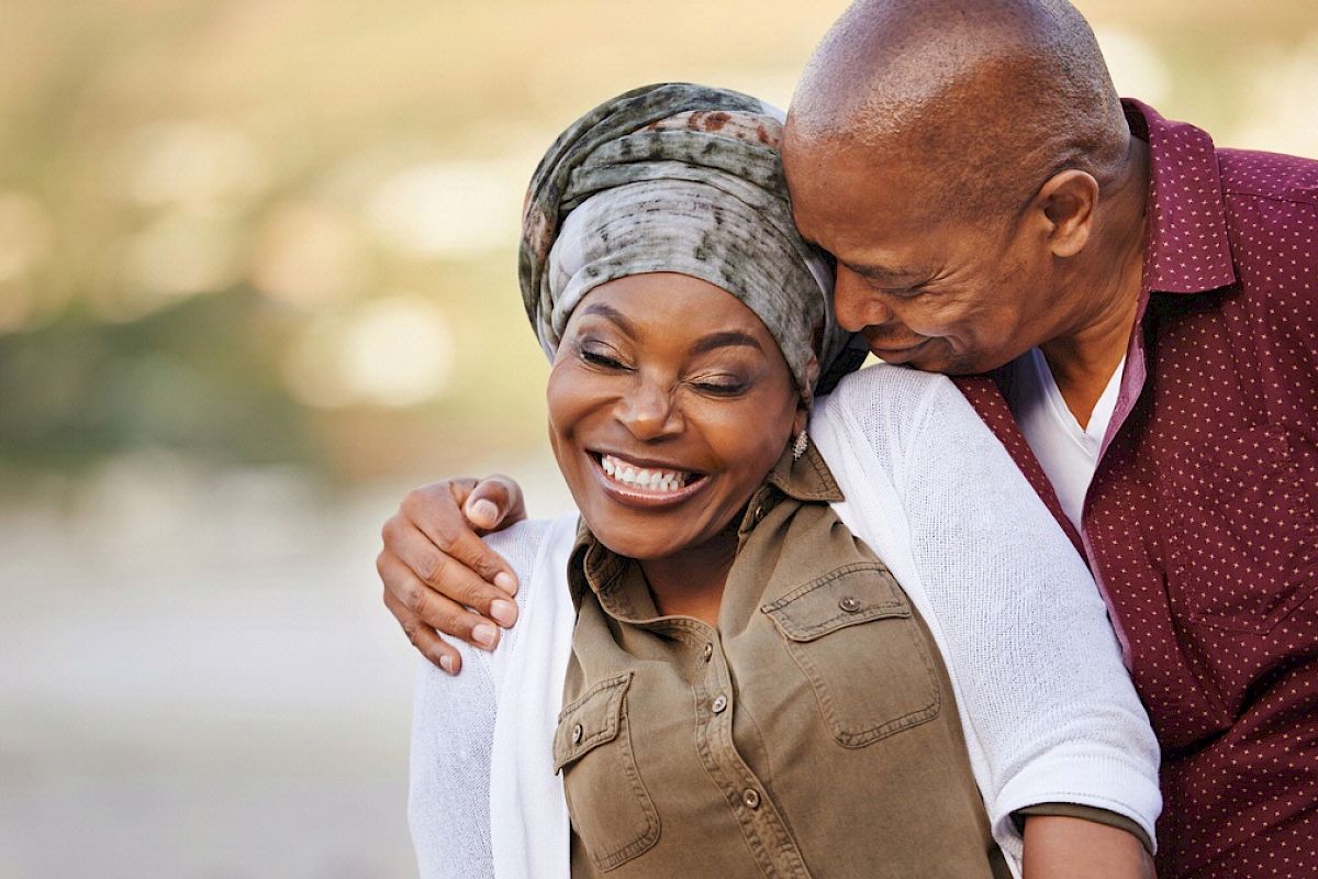 A couple is shown embracing, with the man kissing the woman's head. The woman is smiling and they appear to be outdoors, enjoying a pleasant moment.