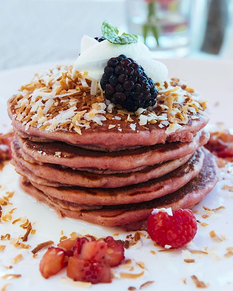 Stack of pink pancakes topped with blueberries, a blackberry, shredded coconut, whipped cream, and fresh berries.