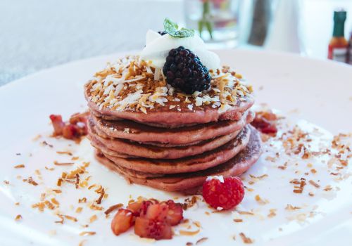 Stack of pink pancakes topped with blueberries, a blackberry, shredded coconut, whipped cream, and fresh berries.