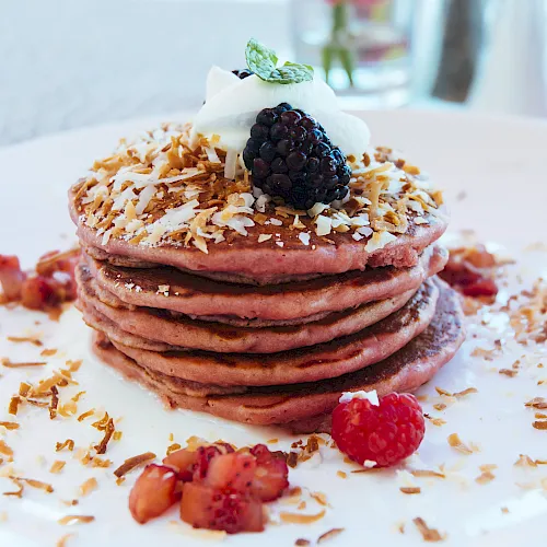 Stack of pink pancakes topped with blueberries, a blackberry, shredded coconut, whipped cream, and fresh berries.