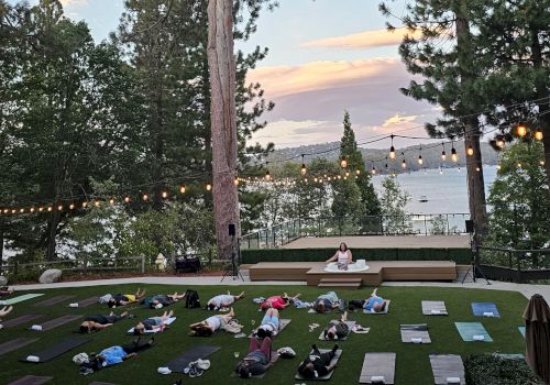 Outdoor yoga in a grassy lawn at sunset, with people on mats, string lights, tall pine trees, and a lake and hills in view.