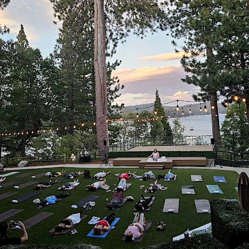 Outdoor yoga in a grassy lawn at sunset, with people on mats, string lights, tall pine trees, and a lake and hills in view.