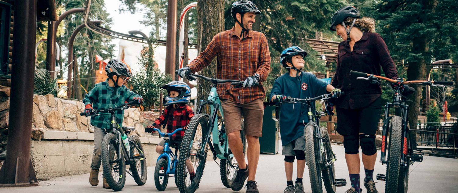 A family in helmets with mountain bikes on a paved path through a forest park.