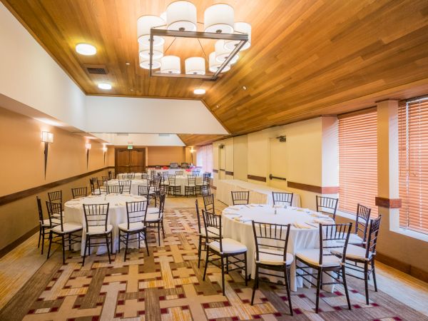 An empty banquet hall with round tables and chairs, patterned carpet, and chandeliers.